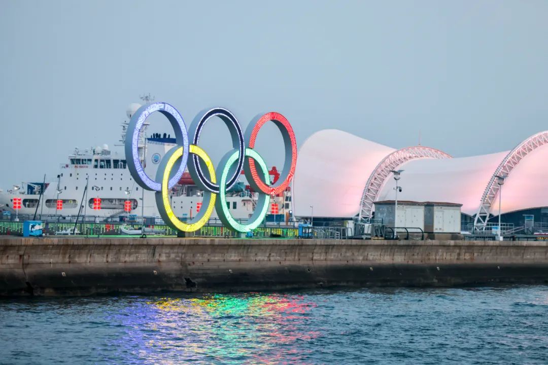 Qingdao Olympic Sailing Centre at dusk — illuminated Olympic rings reflected in the harbour water, with the iconic wave-shaped sailing venue roof in the background, Qingdao, Shandong Province