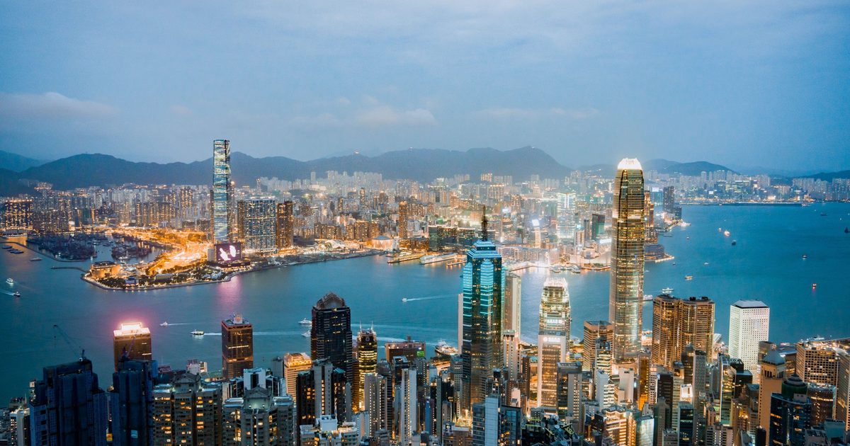 Hong Kong Island towers lit at dusk above Victoria Harbour, with Kowloon’s skyline and hills beyond