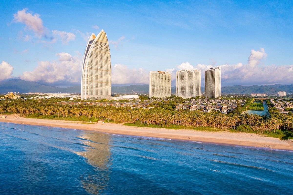 Aerial view of Haitang Bay, Sanya, Hainan — the iconic sail-shaped luxury hotel tower rising above palm-lined white sand beach and turquoise South China Sea, with resort villas and mountain backdrop